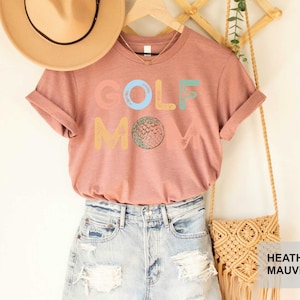 May include: A heather mauve t-shirt with the text "GOLF MOM" in colorful letters. The shirt is styled with a straw hat, denim shorts, and a macrame bag.
