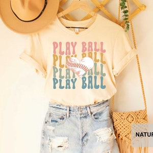 Peut inclure: Un t-shirt beige clair avec un graphique r&eacute;tro d'un ballon de baseball en forme de c&oelig;ur et le texte "PLAY BALL" r&eacute;p&eacute;t&eacute; dans diff&eacute;rentes couleurs. Le t-shirt est port&eacute; par une personne portant un short en jean.