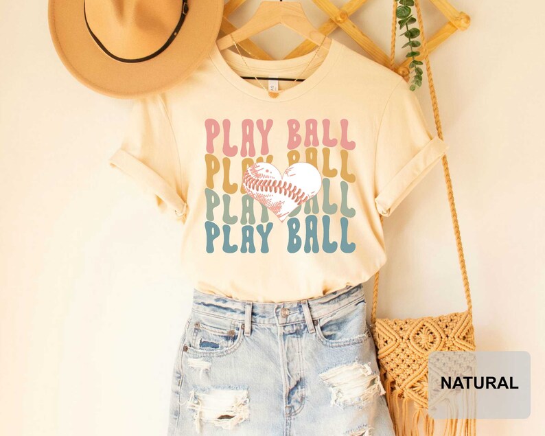Peut inclure: Un t-shirt beige clair avec un graphique r&eacute;tro d'un ballon de baseball en forme de c&oelig;ur et le texte "PLAY BALL" r&eacute;p&eacute;t&eacute; dans diff&eacute;rentes couleurs. Le t-shirt est port&eacute; par une personne portant un short en jean.