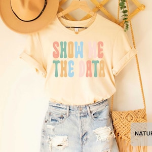 May include: A light beige t-shirt with the text "SHOW ME THE DATA" in colorful retro lettering. The shirt is being modeled with a straw hat and denim shorts.