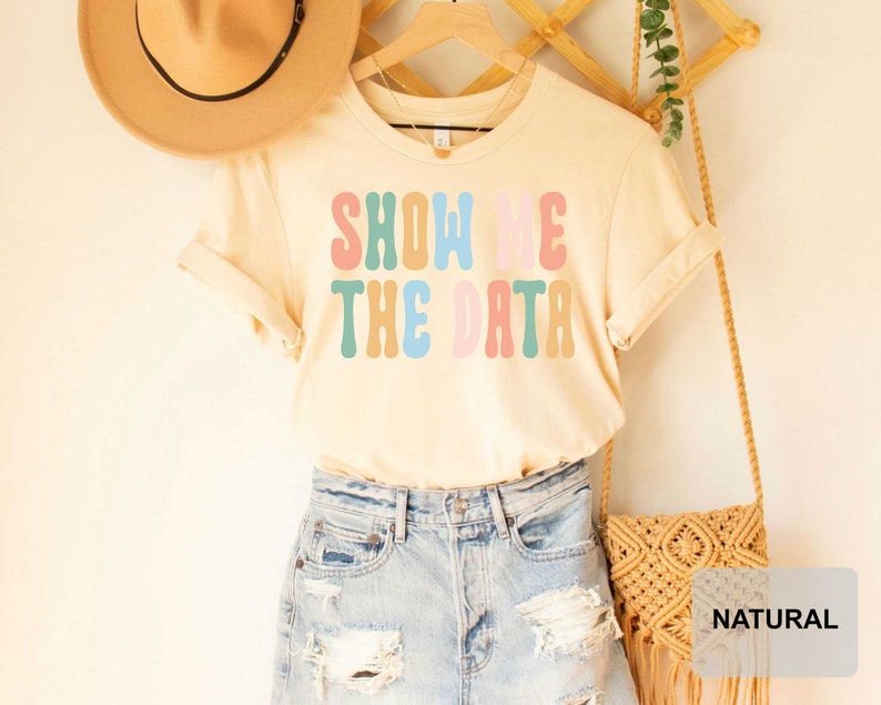 May include: A light beige t-shirt with the text "SHOW ME THE DATA" in colorful retro lettering. The shirt is being modeled with a straw hat and denim shorts.