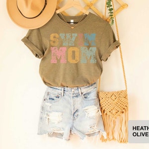 Pu&ograve; includere: Una maglietta verde con la scritta "Swim Mom" in lettere colorate e consumate. La maglietta &egrave; abbinata a pantaloncini di jeans e una borsa all'uncinetto.