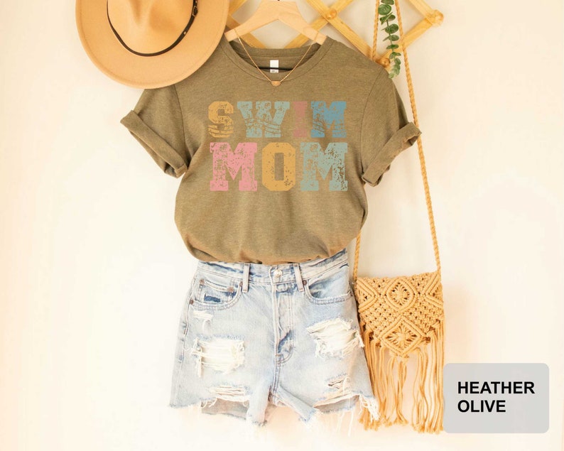 Pu&ograve; includere: Una maglietta verde con la scritta "Swim Mom" in lettere colorate e consumate. La maglietta &egrave; abbinata a pantaloncini di jeans e una borsa all'uncinetto.