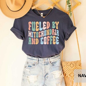 Op de afbeelding: Donkerblauw T-shirt met de tekst "Fueled by Mitochondria and Coffee" in kleurrijke retro letters.