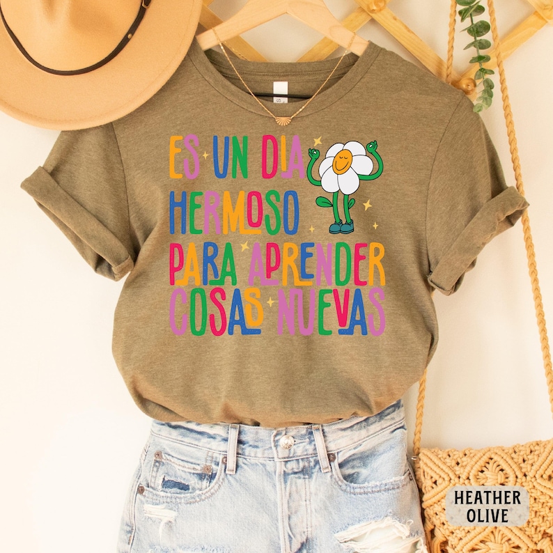 May include: A green t-shirt with the text "Es un dia hermoso para aprender cosas nuevas" in colorful letters. A cartoon flower with a smiling face is in the center of the text.