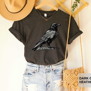 May include: Dark grey heather V-neck t-shirt with a black and white illustration of a raven. The shirt is shown on a model wearing denim shorts and a straw hat.