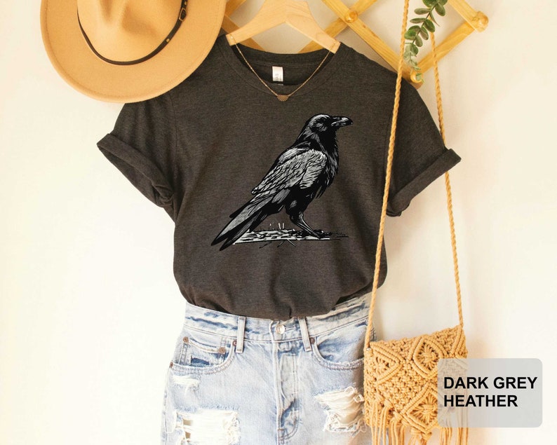 May include: Dark grey heather V-neck t-shirt with a black and white illustration of a raven. The shirt is shown on a model wearing denim shorts and a straw hat.