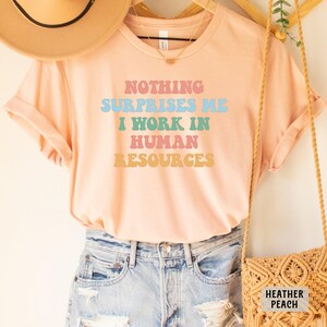 May include: A peach colored t-shirt with the text "Nothing Surprises Me I Work In Human Resources" in rainbow colors. The shirt is on a model wearing denim shorts.