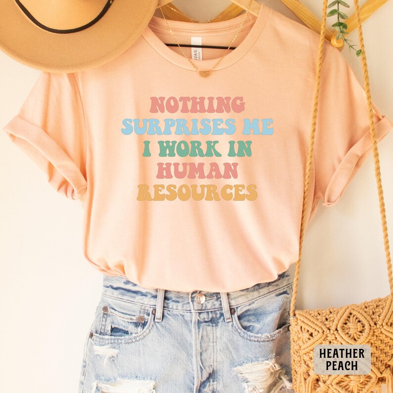 May include: A peach colored t-shirt with the text "Nothing Surprises Me I Work In Human Resources" in rainbow colors. The shirt is on a model wearing denim shorts.