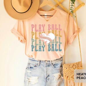 Peut inclure: Un t-shirt couleur p&ecirc;che avec le texte "PLAY BALL" r&eacute;p&eacute;t&eacute; quatre fois dans une police r&eacute;tro. Un ballon de baseball en forme de c&oelig;ur se trouve au centre du texte. Le t-shirt est port&eacute; par une personne portant un short en jean et un chapeau de paille.