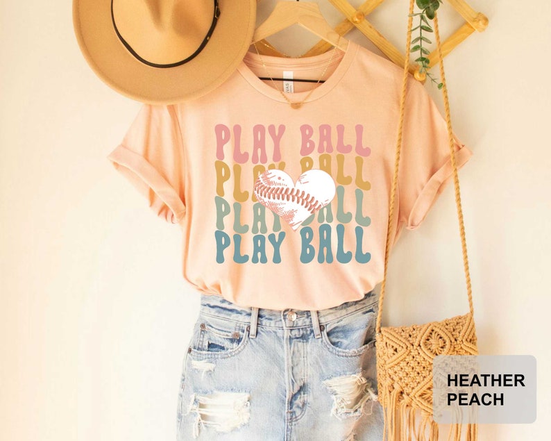 Peut inclure: Un t-shirt couleur p&ecirc;che avec le texte "PLAY BALL" r&eacute;p&eacute;t&eacute; quatre fois dans une police r&eacute;tro. Un ballon de baseball en forme de c&oelig;ur se trouve au centre du texte. Le t-shirt est port&eacute; par une personne portant un short en jean et un chapeau de paille.