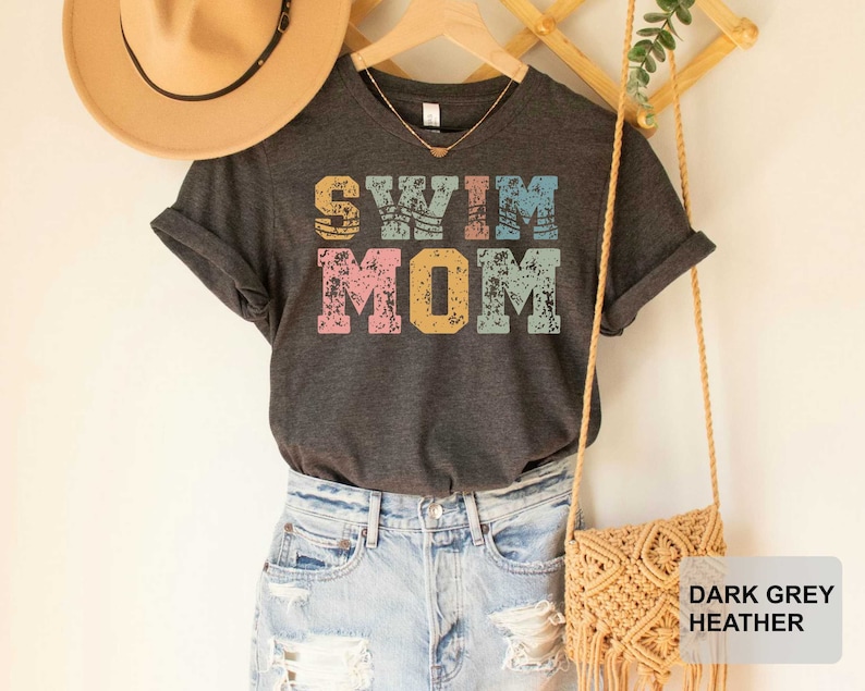 Pu&ograve; includere: Maglietta in grigio melange scuro con il testo "Swim Mom" in lettere colorate e invecchiate. La maglietta &egrave; mostrata su un manichino che indossa pantaloncini di jeans e un cappello di paglia.