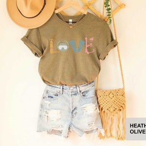 Peut inclure: Un t-shirt en couleur olive chin&eacute; avec un motif repr&eacute;sentant le mot "LOVE" &eacute;crit avec du mat&eacute;riel de plong&eacute;e sous-marine. Le t-shirt est associ&eacute; &agrave; un short en jean et un sac en paille.