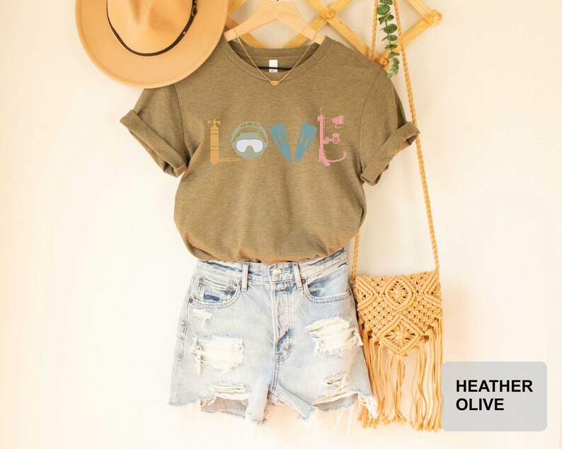 Peut inclure: Un t-shirt en couleur olive chin&eacute; avec un motif repr&eacute;sentant le mot "LOVE" &eacute;crit avec du mat&eacute;riel de plong&eacute;e sous-marine. Le t-shirt est associ&eacute; &agrave; un short en jean et un sac en paille.