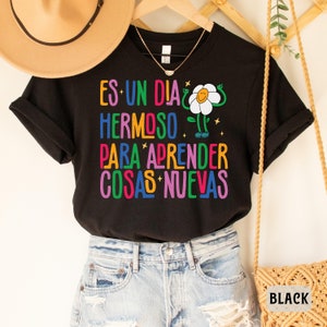 May include: Black t-shirt with a colorful graphic design featuring a flower with a smiling face and the text "Es un dia hermoso para aprender cosas nuevas".
