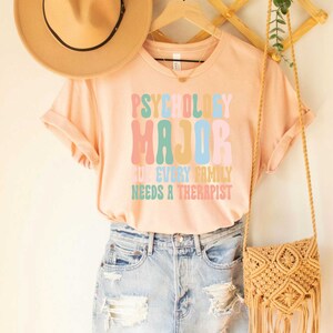 May include: A peach-colored t-shirt with the text "Psychology Major" in rainbow colors. The text below reads "Cuz every family needs a therapist".