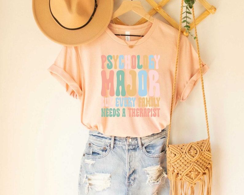 May include: A peach-colored t-shirt with the text "Psychology Major" in rainbow colors. The text below reads "Cuz every family needs a therapist".