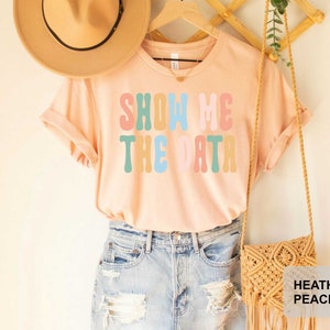 May include: A light pink t-shirt with the text "Show Me The Data" in colorful retro font. The shirt is worn with blue denim shorts and a straw bag.
