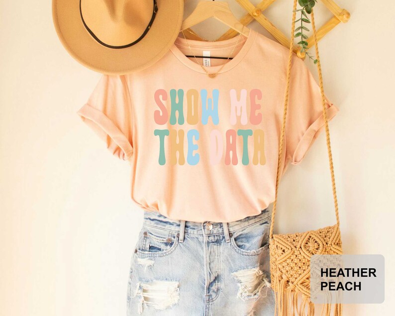 May include: A light pink t-shirt with the text "Show Me The Data" in colorful retro font. The shirt is worn with blue denim shorts and a straw bag.