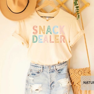 Puede incluir: Una camiseta beige claro con el texto "SNACK DEALER" en letras de colores. La camiseta se muestra en una percha con un sombrero de paja y una bolsa tejida.
