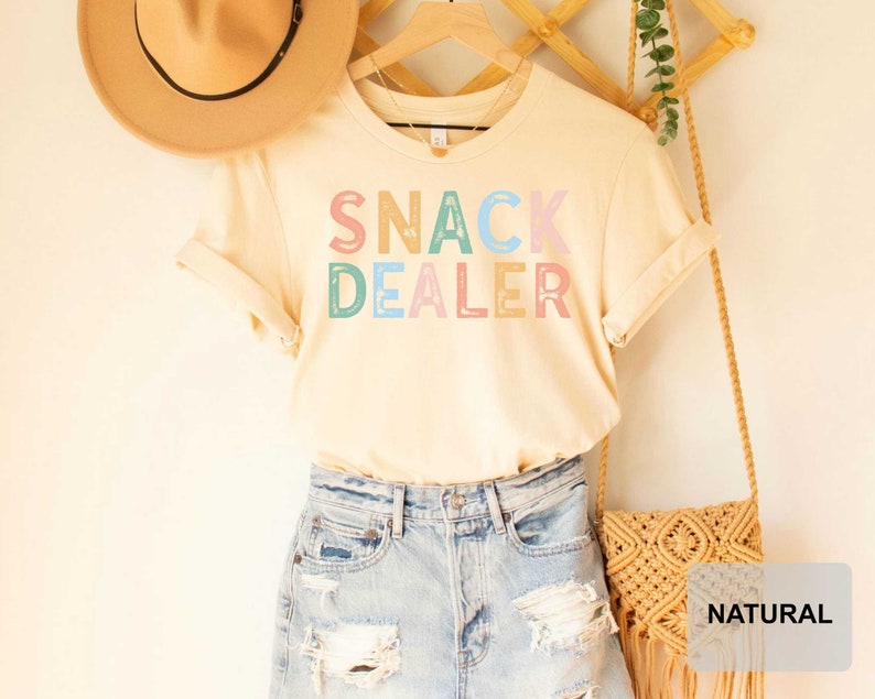 Puede incluir: Una camiseta beige claro con el texto "SNACK DEALER" en letras de colores. La camiseta se muestra en una percha con un sombrero de paja y una bolsa tejida.