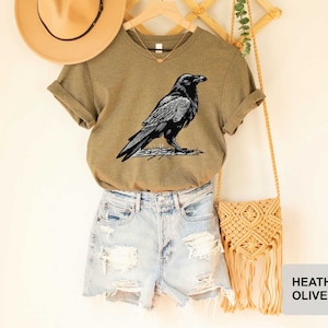 May include: A heather olive t-shirt featuring a black raven illustration. The t-shirt is paired with denim shorts and a macrame bag.