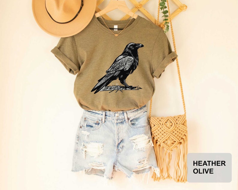 May include: A heather olive t-shirt featuring a black raven illustration. The t-shirt is paired with denim shorts and a macrame bag.