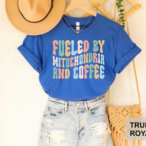 Op de afbeelding: Een koninklijk blauw T-shirt met de tekst "Fueled by Mitochondria and Coffee" in een kleurrijk retro-lettertype.