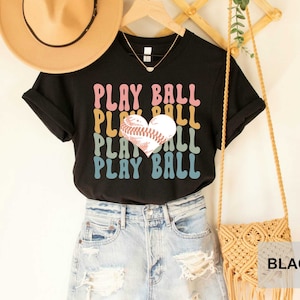 Peut inclure: T-shirt noir avec un graphique r&eacute;tro d'un ballon de baseball en forme de c&oelig;ur et le texte "PLAY BALL" r&eacute;p&eacute;t&eacute; dans diff&eacute;rentes couleurs.