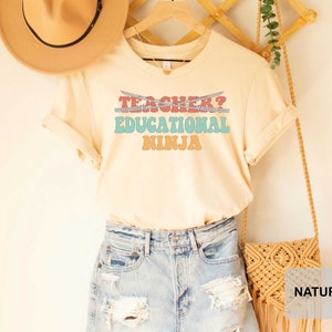 May include: A light beige t-shirt with the text "Teacher? Educational Ninja" in a retro font with colorful accents. The shirt is displayed on a mannequin with a straw hat and a denim skirt.