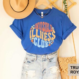 K&ouml;nnte beinhalten: Blaues T-Shirt mit dem Text "Invisible Illness Club" in einer Retro-Schriftart mit bunten Blumen, die den Text umgeben. Das Shirt wird mit einer Jeans-Shorts und einer Strohtasche modelliert.