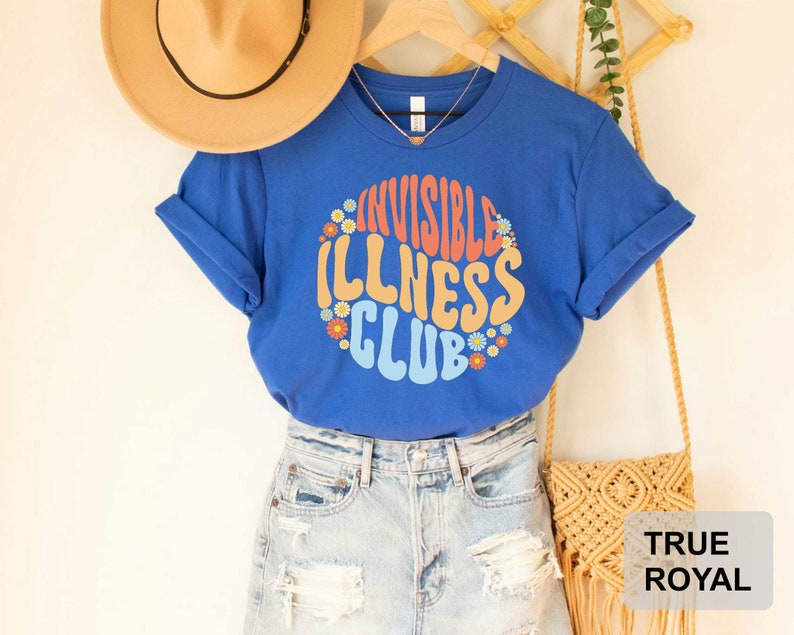 K&ouml;nnte beinhalten: Blaues T-Shirt mit dem Text "Invisible Illness Club" in einer Retro-Schriftart mit bunten Blumen, die den Text umgeben. Das Shirt wird mit einer Jeans-Shorts und einer Strohtasche modelliert.