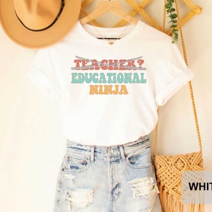 May include: White t-shirt with the text "Teacher? Educational Ninja" in a retro font with colorful accents. The shirt is being modeled with a pair of denim shorts and a straw hat.