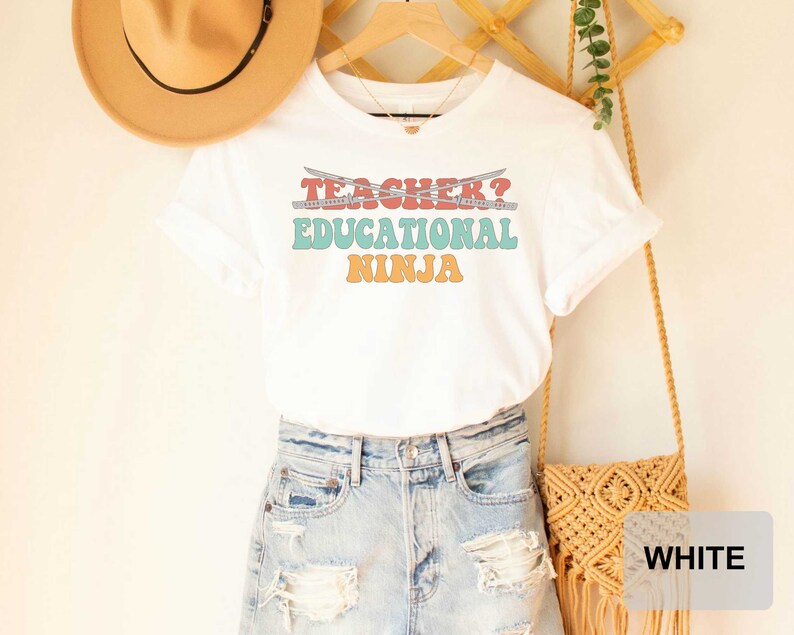 May include: White t-shirt with the text "Teacher? Educational Ninja" in a retro font with colorful accents. The shirt is being modeled with a pair of denim shorts and a straw hat.