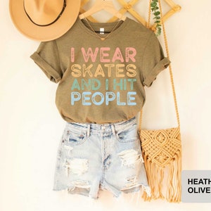 May include: A heather olive green t-shirt with the text "I wear skates and I hit people" in colourful letters. The shirt is modelled on a person wearing denim shorts.