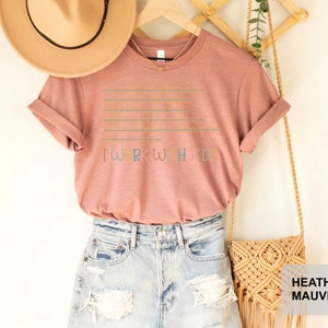 May include: A heather mauve t-shirt with the text "I work with my hands" in colorful letters. The shirt is displayed on a hanger with a straw hat and a macrame bag.