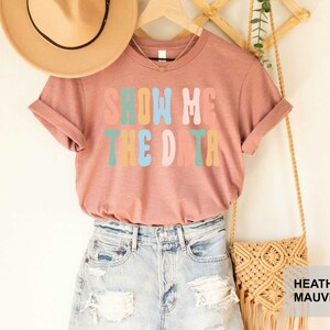 May include: A heather mauve t-shirt with the text "Show Me The Data" in rainbow colors. The shirt is displayed on a model with a straw hat and a crocheted bag.
