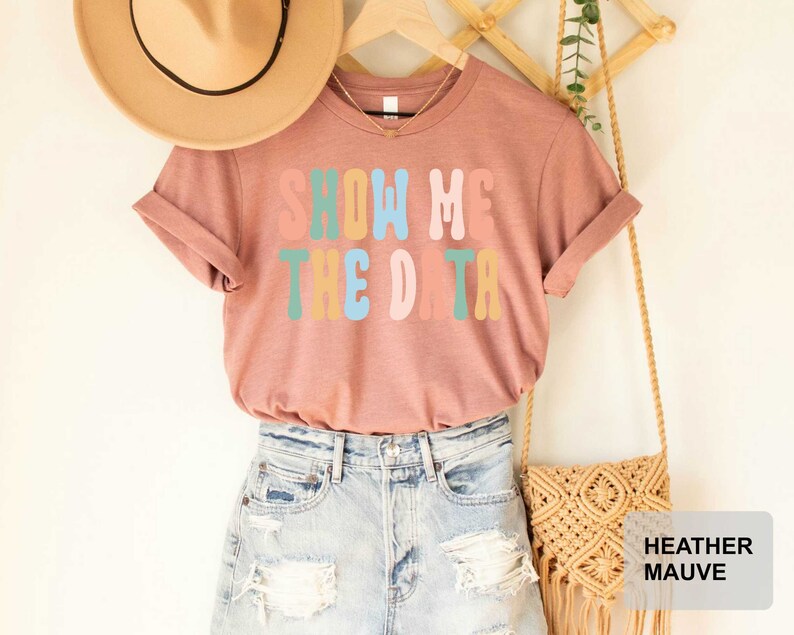 May include: A heather mauve t-shirt with the text "Show Me The Data" in rainbow colors. The shirt is displayed on a model with a straw hat and a crocheted bag.