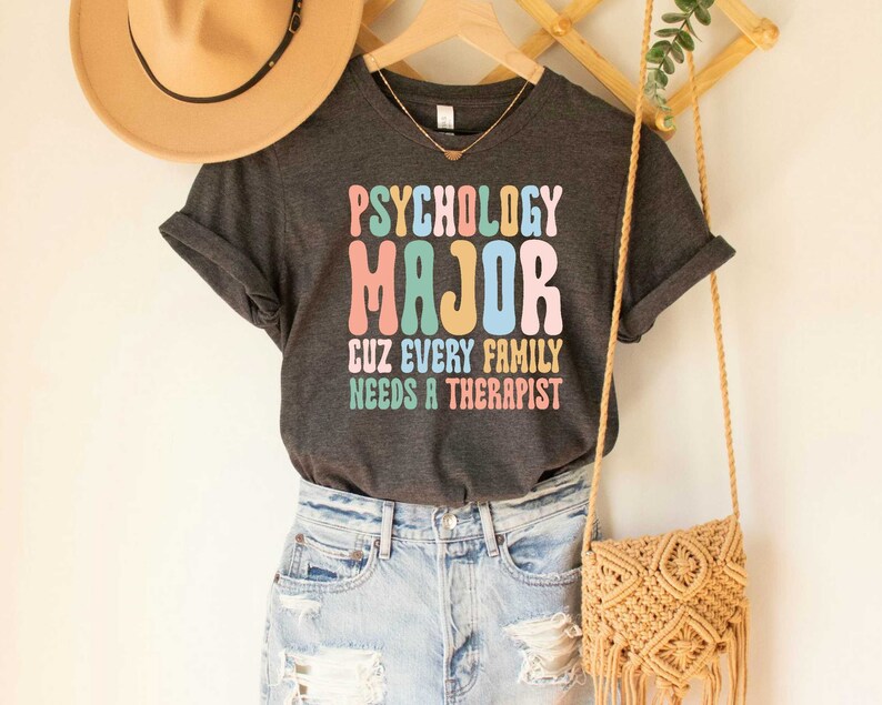 May include: A dark grey t-shirt with the text "Psychology Major Cuz Every Family Needs A Therapist" in colorful, retro-style lettering.