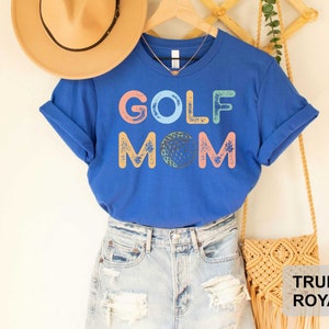 May include: A royal blue t-shirt with the text "GOLF MOM" in colorful letters with a golf ball graphic. The shirt is being modeled with a pair of light blue denim jeans and a straw hat.