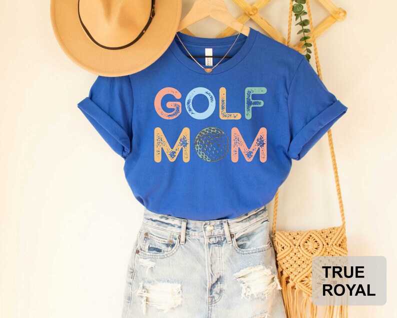May include: A royal blue t-shirt with the text "GOLF MOM" in colorful letters with a golf ball graphic. The shirt is being modeled with a pair of light blue denim jeans and a straw hat.