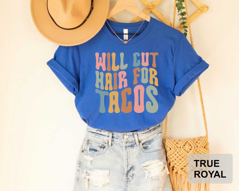 May include: A blue t-shirt with the text "Will Cut Hair For Tacos" in colorful, retro-style lettering. The shirt is being modeled on a person wearing denim shorts and a straw hat.