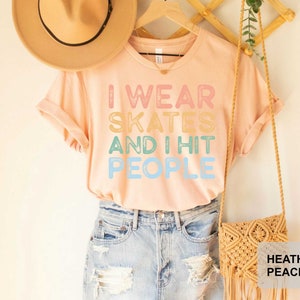 May include: A light pink t-shirt with the text "I wear skates and I hit people" in rainbow colours. The shirt is being modelled by a person wearing blue denim shorts and a brown hat.