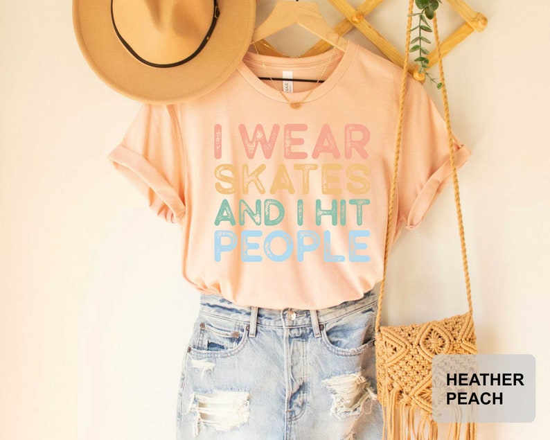 May include: A light pink t-shirt with the text "I wear skates and I hit people" in rainbow colours. The shirt is being modelled by a person wearing blue denim shorts and a brown hat.