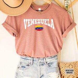 May include: A heather mauve t-shirt with the word "VENEZUELA" in white text with a distressed look. The Venezuelan flag is printed below the text.