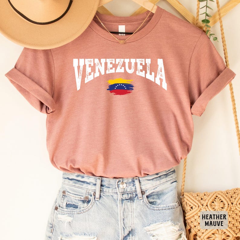 May include: A heather mauve t-shirt with the word "VENEZUELA" in white text with a distressed look. The Venezuelan flag is printed below the text.