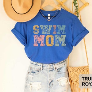 Pu&ograve; includere: Una maglietta blu reale con il testo "Swim Mom" in un font in stile vintage con un aspetto usurato. Il testo &egrave; in tonalit&agrave; di rosa, giallo e blu.