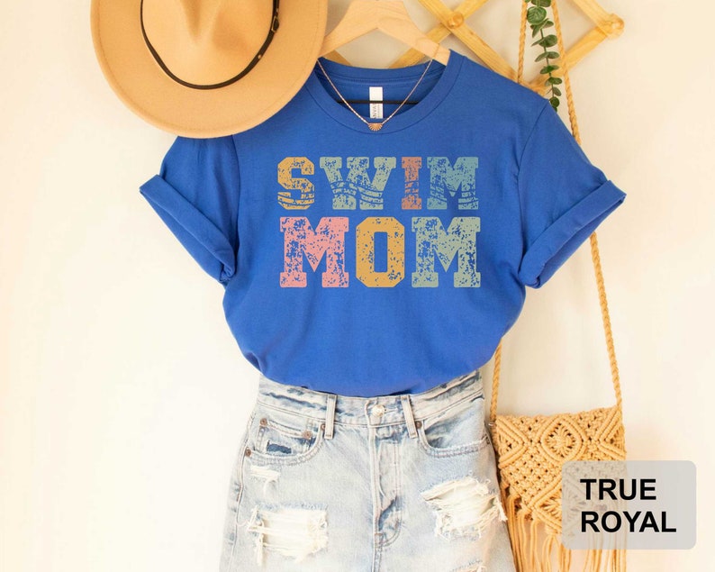 Pu&ograve; includere: Una maglietta blu reale con il testo "Swim Mom" in un font in stile vintage con un aspetto usurato. Il testo &egrave; in tonalit&agrave; di rosa, giallo e blu.