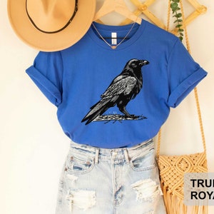 May include: A royal blue t-shirt with a black and white illustration of a raven. The t-shirt has a crew neck and short sleeves.