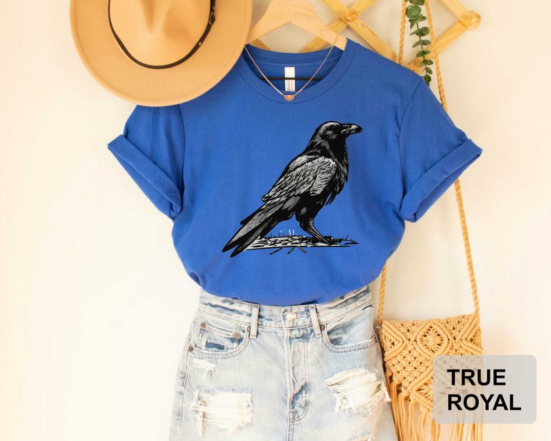 May include: A royal blue t-shirt with a black and white illustration of a raven. The t-shirt has a crew neck and short sleeves.